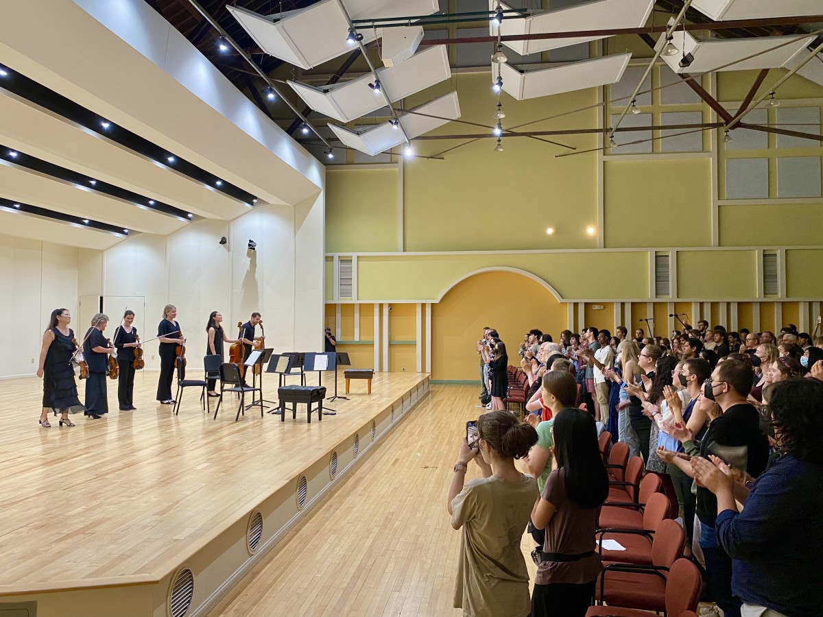 A side view of a concert stage with musicians bowing on the left and audience members applauding on the right