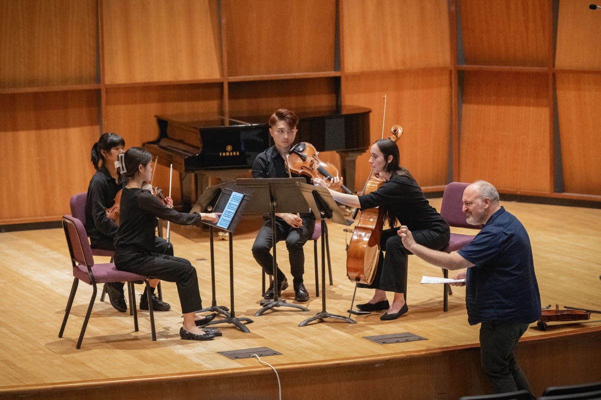 A string quartet of students sitting on stage while a guest musician stands in front of them and gives musical suggestions