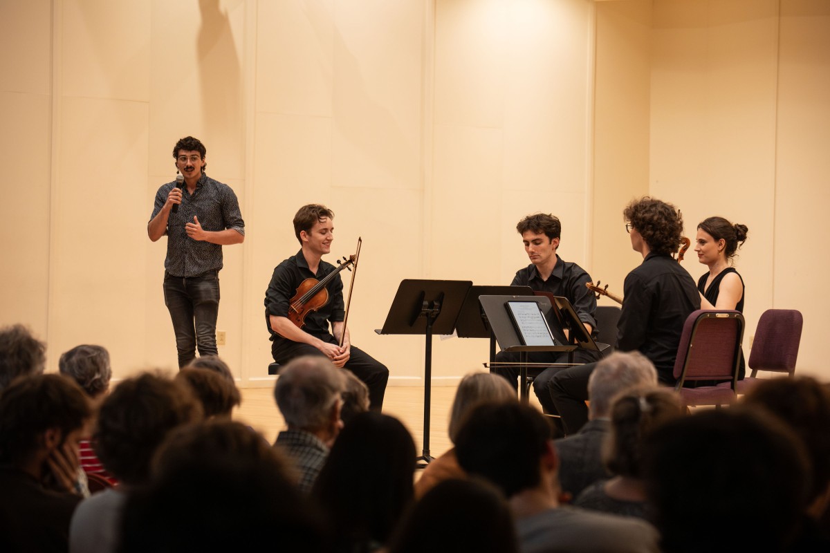 Four musicians sitting on stage while a fifth, the composer of the work, speaks from stage into a microphone
