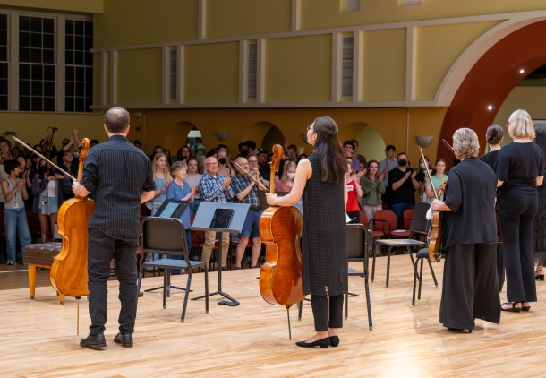 A view from behind of string musicians standing on stage with their instruments facing an audience which is giving a standing ovation.