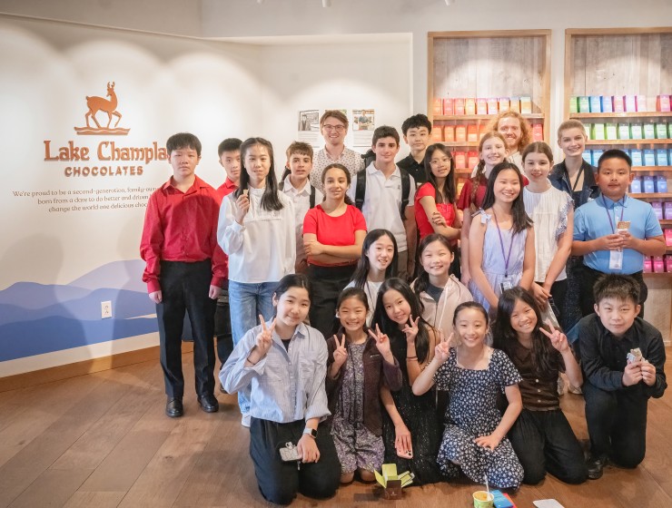 a group of young sudents posing inside Lake Champlain Chocolates; the logo and name of the business are on the wall behind the,