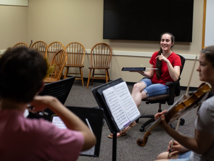 A festival fellow wearing a red shirt sitting in a chair in front of a student string quartet giving instruction