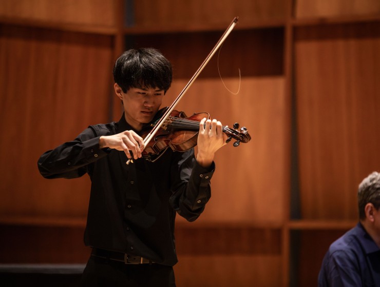 a student violinist performing a solo on stage with a pianist partially in the background