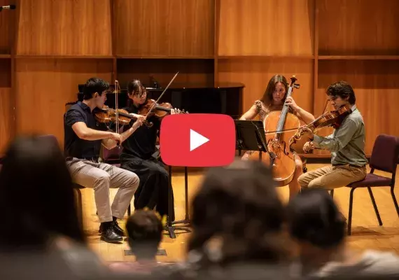 Students performing on stage in a string quartet with a red 'play' button overtop, indicating a YouTube video